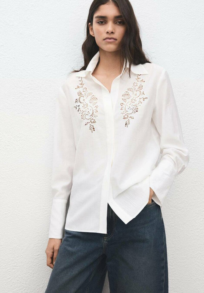 Camisa blanca de manga larga con detallado encaje en la parte frontal, diseño abotonado y corte relajado, combinada con jeans azul oscuro.