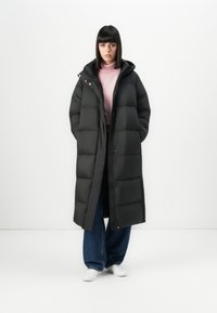 Donna che indossa un lungo cappotto imbottito nero sopra un dolcevita rosa, jeans a gamba larga e sneakers bianche, in piedi contro uno sfondo bianco semplice.