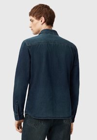 Tmavě modrá džínová košile s dlouhými rukávy, knoflíkovými manžetami a klasickým límcem. Má akcenty v podobě prošití a rovný lem.