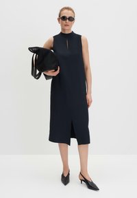 Marineblaues ärmelloses Kleid mit hohem Ausschnitt, Schlitz vorne und A-Linienform. Kombiniert mit einer strukturierten schwarzen Tasche und schwarzen spitzen Absätzen.