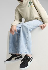 Felpa con cappuccio color beige chiaro con logo grafico, abbinata a jeans di denim azzurro chiaro a gamba larga e sneakers nere in suede con dettagli bianchi.