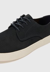 Zapatos de ante negro con una textura suave, que presentan cordones grises, suelas de goma blanca y un sutil hilo azul a lo largo de los bordes.