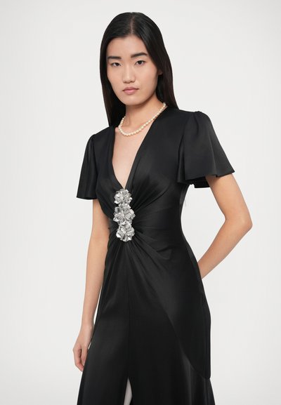 Fekete szatén ruha rövid, fodros ujjakkal, mély V-nyakkivágással és ezüst virágdísszel a deréknál. Oldalsó sliccel rendelkezik.