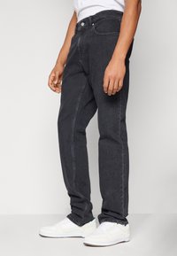 Jeans de mezclilla negra con un corte recto, costuras visibles y un diseño estándar de cinco bolsillos, combinados con zapatillas blancas.