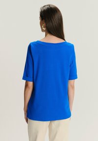 Kobieta z długimi brązowymi włosami, nosząca złote kolczyki w kształcie kół, jasnoniebieską bluzkę z krótkim rękawem i beżowe spodnie, znajdująca się tyłem na tle jednolitego tła.