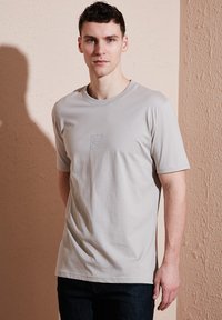 Světle šedé tričko s krátkým rukávem a kulatým výstřihem, s raženým logem na hrudi, vyrobené z měkké bavlny. Přiléhavý střih.
