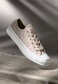 Sneakers na sznurówki z teksturowanym, beżowym nałożeniem z koronki na jasnej podeszwie. Posiadają białą gumową osłonę na palce oraz dopasowane oczka i sznurówki.