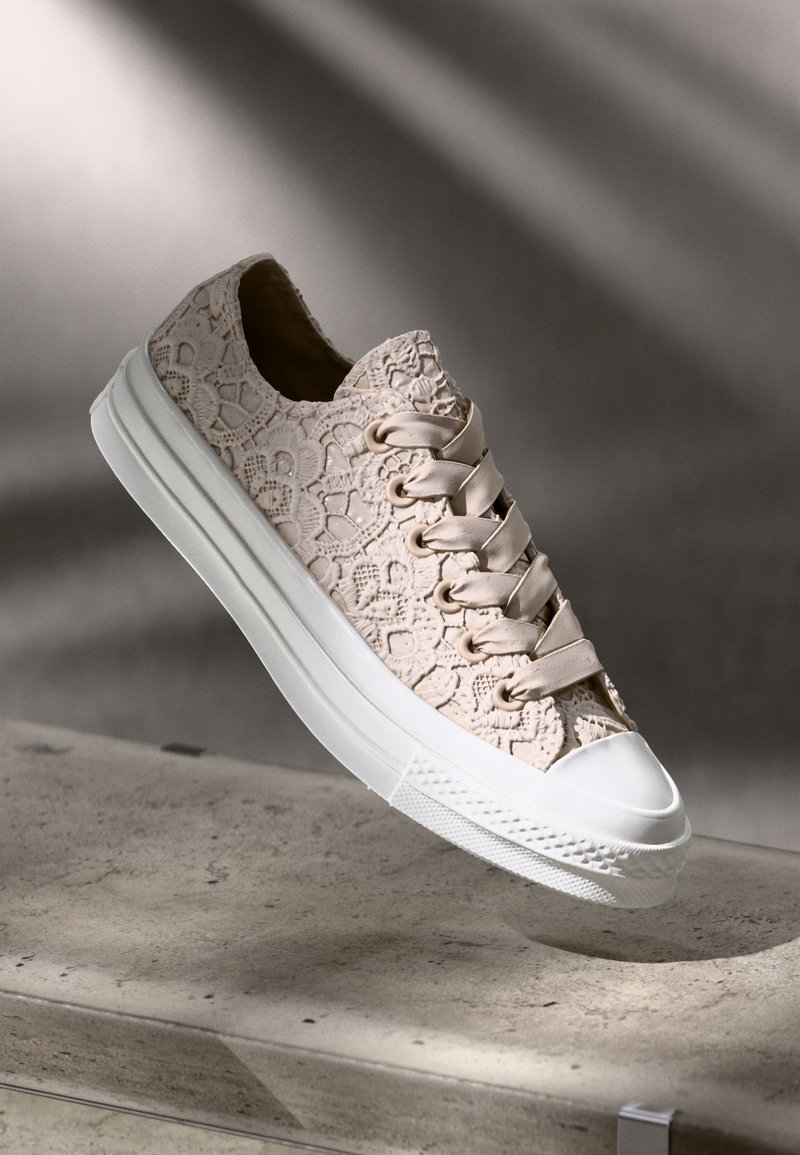 Sneakers na sznurówki z teksturowanym, beżowym nałożeniem z koronki na jasnej podeszwie. Posiadają białą gumową osłonę na palce oraz dopasowane oczka i sznurówki.