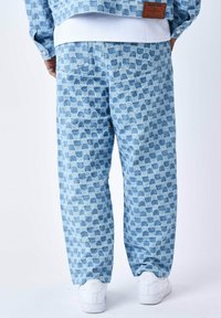 Pantaloni blu a fantasia realizzati in tessuto testurizzato, con un design astratto a quadrati in diverse tonalità di blu e bianco. Accentuati con scarpe bianche.