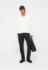 Mann mit schwarzer Mütze, weißem langärmligem Polohemd, schwarzen Hosen, schwarzen Schuhen, der eine schwarze Ledertasche hält und vor einem weißen Hintergrund steht.