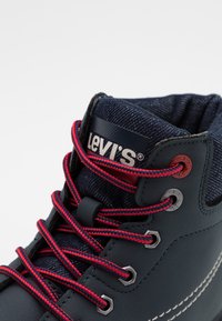 Gros plan sur une chaussure montante Levi's bleu marine avec des lacets rouges et noirs, des œillets métalliques, une languette en tissu et le logo de la marque.