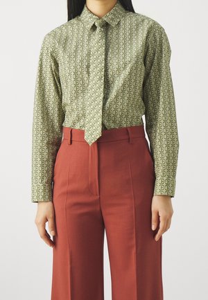 Kobieta w zielonej wzorzystej koszuli z długim rękawem wpuszczonej w szerokie, wysokie, rdzawo-brązowe spodnie na białym tle.