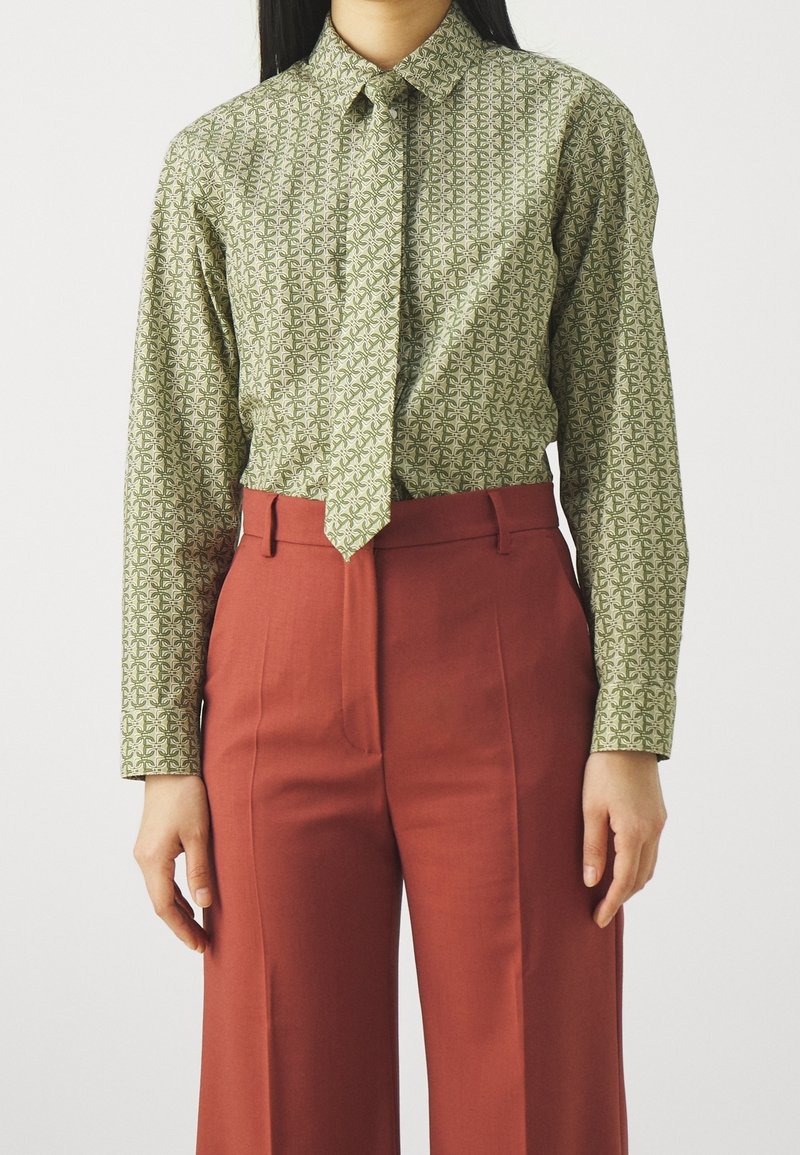 Femme portant une chemise à manches longues verte à motifs rentrée dans un pantalon large taille haute couleur rouille sur fond blanc.
