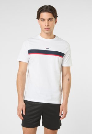 Młody mężczyzna z krótkimi ciemnymi włosami, ubrany w biały t-shirt FILA z granatowo-czerwoną paską oraz czarne spodenki, na jednolitym tle.