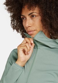 Veste zippée en vert menthe avec un col haut, design légèrement ajusté et un tissu texturé. Le modèle ajuste la fermeture éclair d'une main.