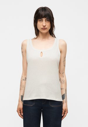 Γυναίκα με σκούρα μαλλιά και τατουάζ στα χέρια, φοράει λευκό αμάνικο τοπ με μικρό άνοιγμα μπροστά και σκούρο μπλε τζιν.