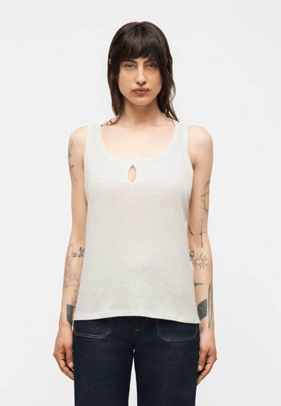 Femme aux cheveux foncés et tatouages sur les bras, portant un haut blanc sans manches avec une petite ouverture sur le devant et un jean bleu foncé.
