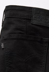 Jean en denim noir avec une finition texturée, comprenant des poches arrière, des coutures incurvées, et un patch en cuir à la taille.