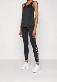 Top sportivo nero senza maniche con texture a rete e bordi in contrasto, abbinato a leggings neri con la scritta "ACTIVE" stampata in colori sfumati. Sneakers bianche.