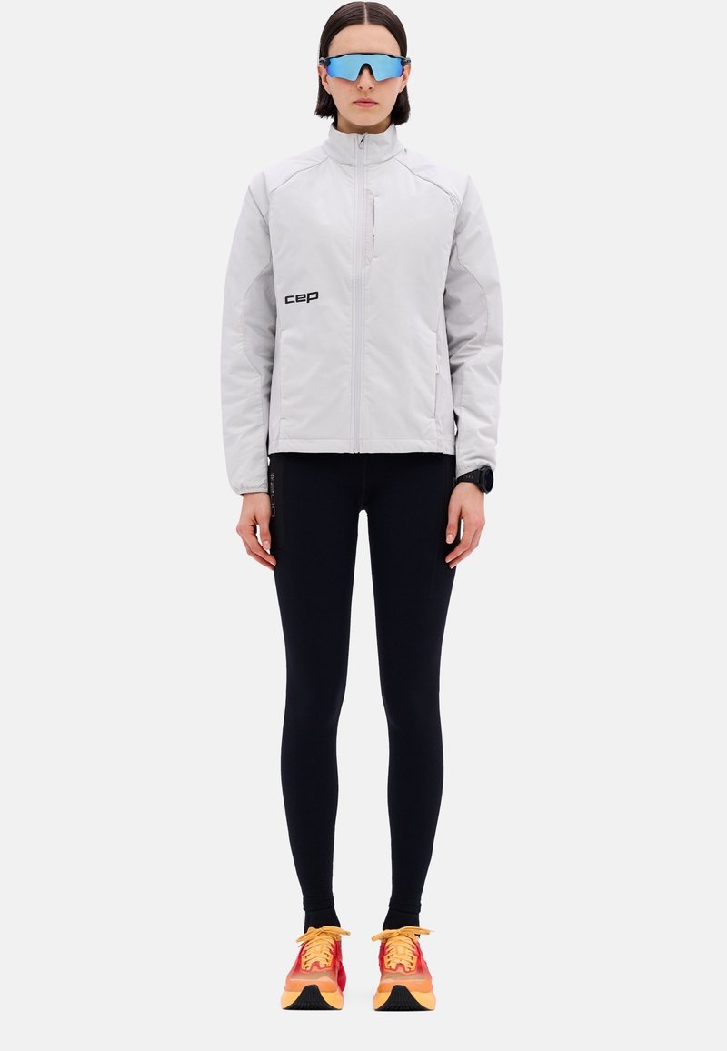 Hellgraue Windjacke mit schwarzem Logo, kombiniert mit schwarzen Leggings und knalligen rot-orangefarbenen Laufschuhen mit gelben Schnürsenkeln.