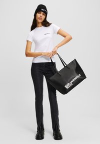 Crna kožna torba s bijelim natpisom "Karl Lagerfeld Jeans", nošena uz bijelu košulju kratkih rukava i crne traperice visokog struka s bočnim prorezima.