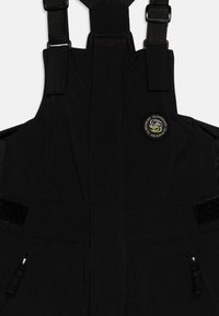 Svart vattentät overall med justerbara remmar, flera blixtlåsfickor och kardborrefickor, med en rund logopatch på bröstet.