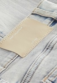 Svjetlosivi traper s bež kožnom etiketom, utisnutom s natpisima "NEXT COLLECTION" i "EST 1962", vidljivim šavovima na rubovima.