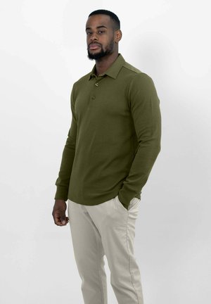 Mann mit kurzem Bart, der ein olivgrünes Langarm-Poloshirt und hellbeige Hosen trägt und mit einer Hand in der Tasche steht.