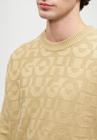 Μπεζ πουλόβερ με ανάγλυφα υφασμάτινα μοτίβα που απεικονίζουν τη λέξη "HUGO" σε διάφορα μεγέθη, στρογγυλή λαιμόκοψη και χερουλάκια με ριμπ.