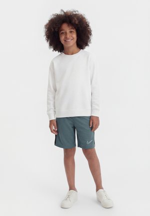 Uśmiechnięte dziecko o kręconych włosach, ubrane w białą bluzę, turkusowe spodenki Nike i białe trampki, stojące na tle białej, jednolitej ściany.