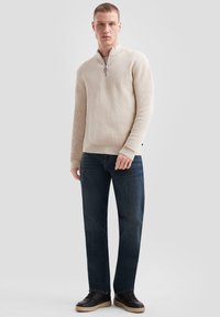 Lichtbeige zipper-trui met ribbeltextuur, gecombineerd met donkerblauwe jeans en zwarte sneakers met een beige zool, staande tegen een grijze achtergrond.
