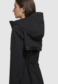 Black waterproof jacket with a cinched waist, a hood, and logo detailing. Smooth texture and clean design with button accents on sleeves.