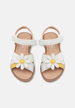 Sandalias blancas para niños con tiras en el tobillo y decoraciones frontales de flores con pétalos blancos y centros amarillos, en una suela beige.