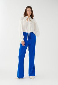 Blusa blanca con cuello lazo y mangas fruncidas combinada con pantalones acampanados de un azul brillante y zapatos de punta beige. Elementos de diseño simples.