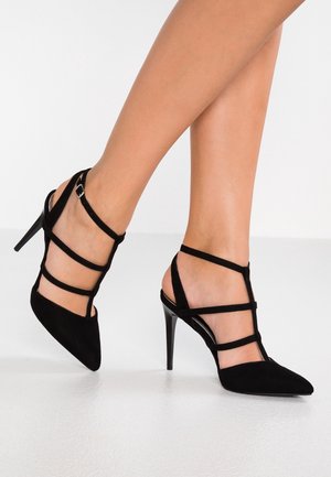 Beine mit schwarzen, spitz zulaufenden High Heels mit mehreren horizontalen und knöchelhohen Riemen vor einem weißen Bodenhintergrund.