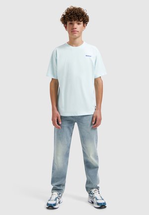 Tiener met krullend haar, gekleed in een lichtblauw oversized T-shirt, verwassen blauwe spijkerbroek en wit-blauwe sneakers, staand tegen een effen achtergrond.