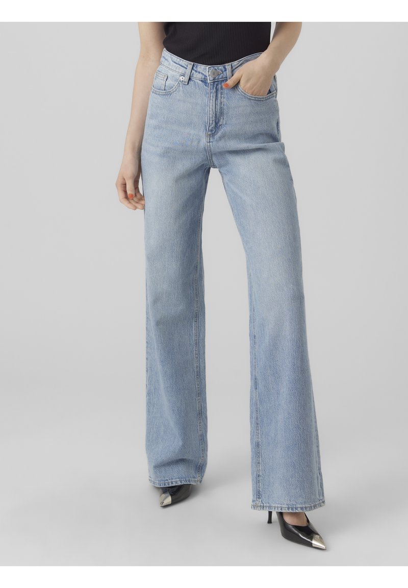 Personne portant un jean large bleu clair et des chaussures à talons hauts noires pointues, debout devant un fond uni.
