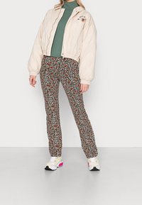 Lichtbeige bomberjack, groene coltrui, gestreepte zwarte broek met kleurrijke bloemen, en witte sneakers met roze accenten.