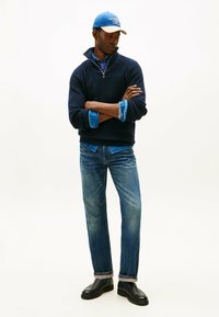 Maglione lavorato a maglia blu navy con colletto a zip, indossato sopra una camicia azzurra, abbinato a jeans azzurri e scarpe nere, completato da un berretto blu.