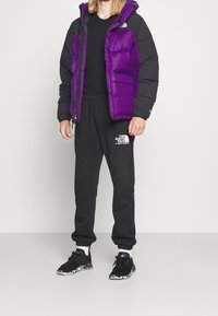 Paarse en zwarte donzen jas met capuchon, ritssluiting en zijzakken; gecombineerd met zwarte joggingbroek en zwarte sneakers met patroonaccenten.