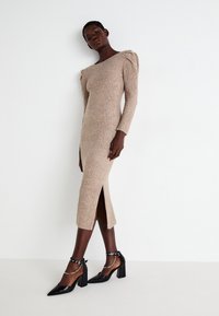 Abito lungo a maglia a coste beige con spacco laterale; abbinato a décolleté nere a punta con cinturini alla caviglia e dettagli metallici.