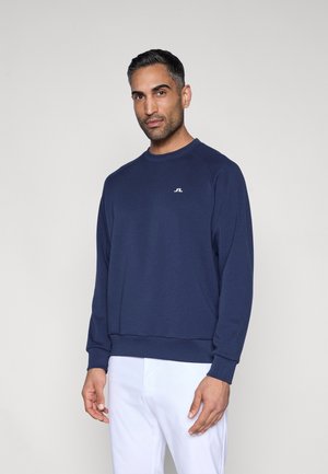 Tengerészkék pulóver puha anyagból, kerek nyakkivágással, bordázott mandzsettákkal és egy kis fehér logóval a mellkasán.