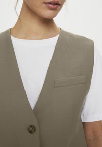 Beige väst med djup V-ringning, en knappstängning och en liten bröstficka. Bärs över en vit T-shirt med en strukturerad tyg.