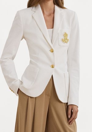 Kvinde iført en tætsiddende hvid blazer med guldknapper og broderet emblem, kombineret med højtaljede beige plisserede bukser.