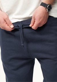 Dunkelblaue Sweatpants mit einem Kordelzug an der Taille, aus einem glatten, strukturierten Stoff. Hände, die den Bund anpassen, sind zu sehen.