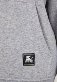 Grijze sweatshirtstof met een zwart-wit Starter Black Label logo patch genaaid nabij de zaknaad.