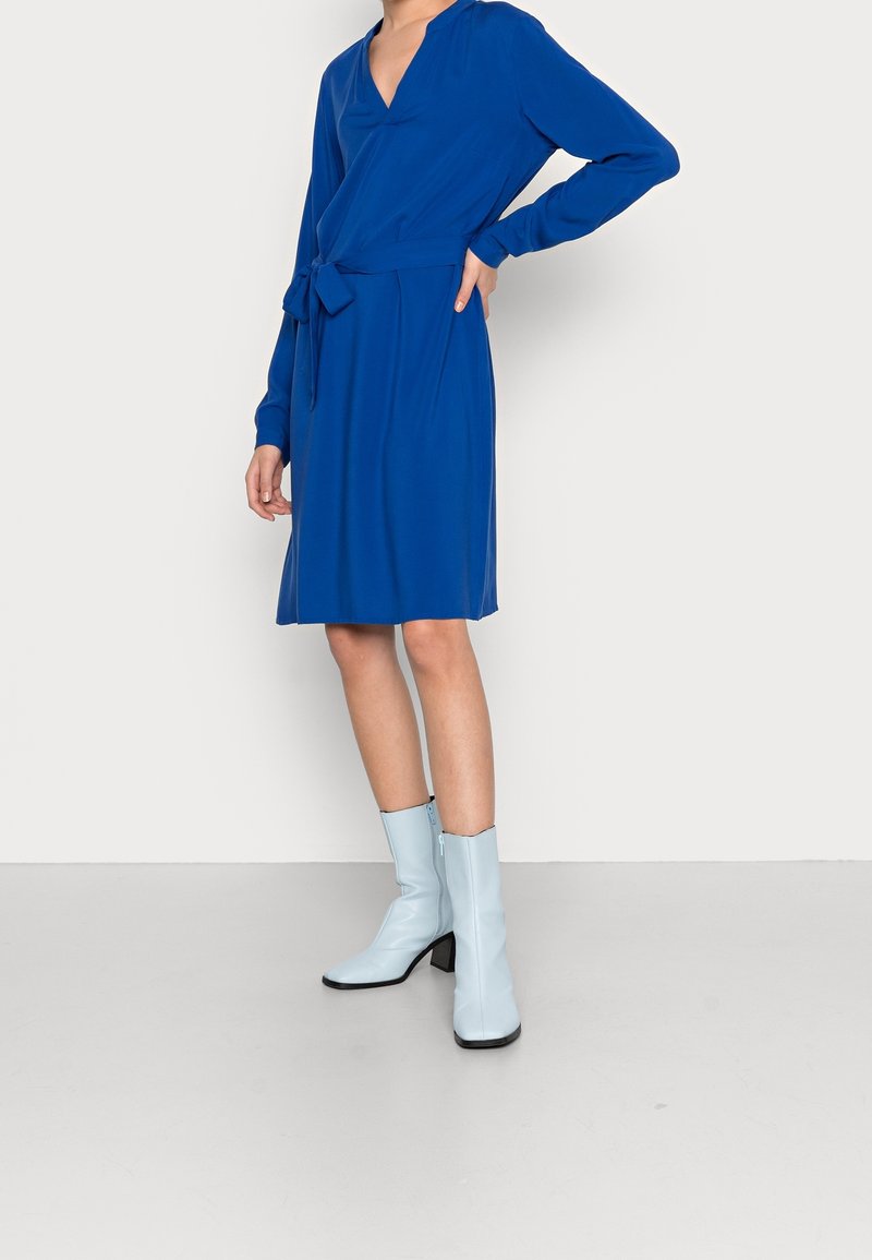 Femme portant une robe bleue mi-longue à manches longues avec une ceinture nouée à la taille, associée à des bottes à talons bleu clair montant jusqu'au milieu du mollet.