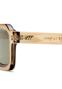 Gros plan sur une branche de lunettes de soleil beige translucide avec charnière métallique et logo "Le Specs" en lettrage doré.