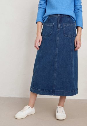 Vrouw in een blauwe sweater gestoken in een hoog-waist denim midi rok met voorzakken, gecombineerd met witte sneakers, staand binnenshuis.