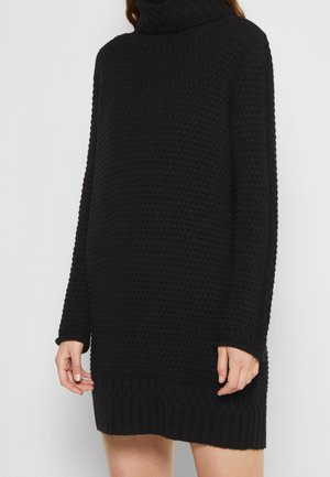 Schwarzes, langärmeliges, gestricktes Rollkragenkleid mit einem strukturierten Muster, getragen von einer Person vor einem einfarbigen hellen Hintergrund.